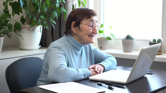 An elderly woman is on a video call, looking at her laptop screen and happily talking. The use of technology for independent living and the importance of online communication for older adults.