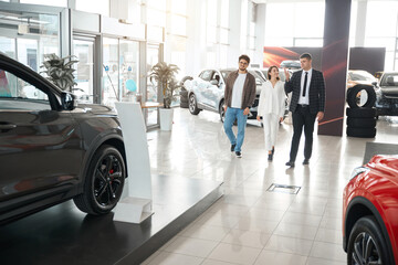 Couple choosing new car with salesman choosing suitable model of auto - obrazy, fototapety, plakaty