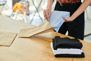 cropped view of skilled woman packing folded clothes in paper bag in print studio, small business