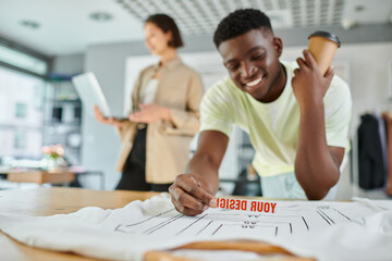 happy african american designer with text template near clothes and asian colleague with laptop