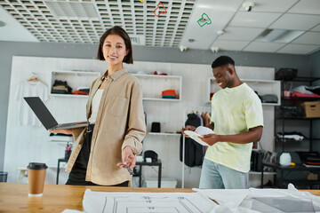 happy asian designer with laptop and text template near african american man in print studio