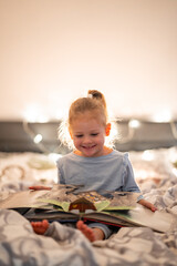 Little girl reading pop up book in home bed in Christmas environment with lights background