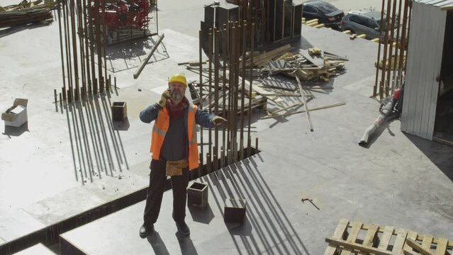 High angle shot of middle-aged Caucasian foreman in workwear using walkie talkie while controlling work of construction crane standing on concrete rooftop of unfinished real estate building