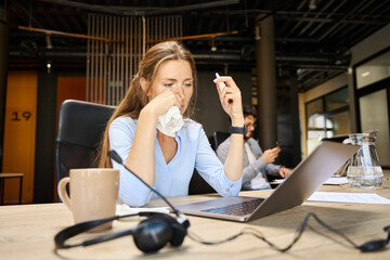 Young sick businesswoman cut off runny nose with napkin during working on laptop