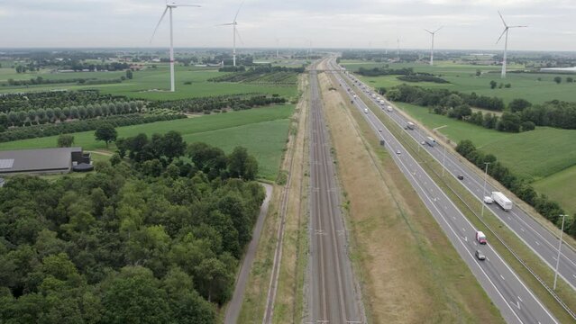 This drone footage captures the E19 highway as it cuts through the scenic landscape of Brecht, Belgium. In the backdrop, wind turbines stand tall, symbolizing a commitment to sustainable energy. The