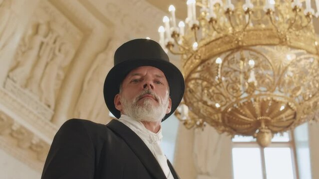 Low angle portrait of grey haired bearded mid aged Caucasian man wearing black tuxedo and cylinder hat looking down at camera standing under luxury golden chandelier in ballroom
