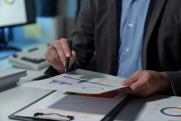 Young Asian businessman holds a pen and points to a chart graph on a document to analyze market data, finance, balance sheet, net profit, taxes to plan an online sales strategy startup business idea.