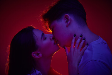 Portrait of young man and woman, loving couple kissing against red background in neon light. Anniversary. Concept of romance, love, relationship, passion, youth, dating, happiness