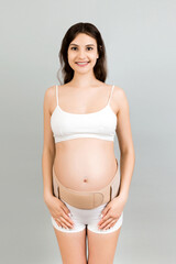 Portrait of pregnant woman in underwear wearing pregnancy bandage at gray background with copy...