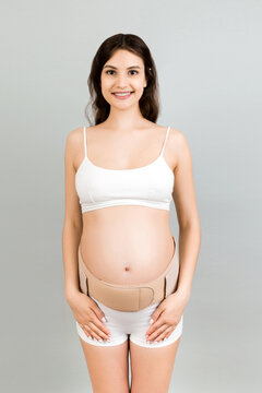 Portrait of pregnant woman in underwear wearing pregnancy bandage at gray background with copy space. Orthopedic abdominal support belt concept