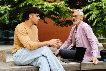 Two male colleagues discussing business project and drinking coffee while sitting outdoors