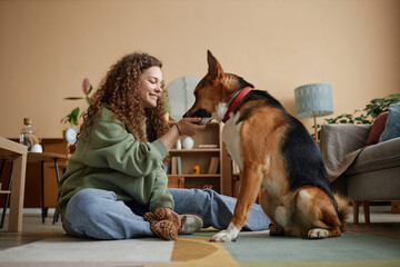 Side view portrait of happy young woman giving treats to pet dog while sitting on floor in cozy...