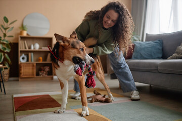 Full length portrait of carefree young woman playing with big dog in cozy living room at home, copy...