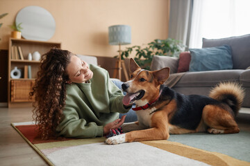 Full length portrait of happy young woman playing with big dog lying on floor in cozy home, copy...