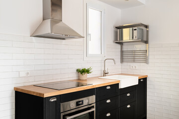 Kitchen island with convection hob and oven with black drawers and wood panel and sink with window and extractor hood against white brick wall. Concept of modern renovation in a new apartment