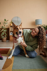 Vertical full length portrait of smiling young woman playing with mixed breed dog on floor in cozy...