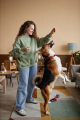 Vertical full length portrait of happy young woman playing with dog jumping for treats in cozy home