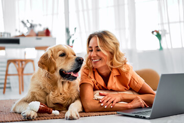 Pet as best friend. Golden retriever resting on floor near smiling female owner using portable laptop. Purebred domestic animal keeping company for charming young woman during video conference.