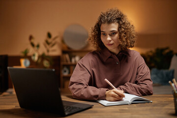 Front view portrait of curly haired young woman using laptop and taking notes while studying online...