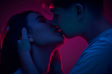 Tender young woman kissing her boyfriend, showing love and care against purple background in neon light. Concept of romance, love, relationship, passion, youth, dating, happiness