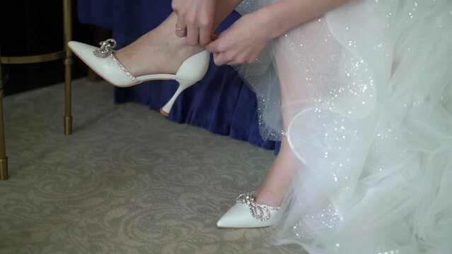 Bride put on wedding shoes. White luxury bridal footwear. Young woman in long luxury bridal gown.