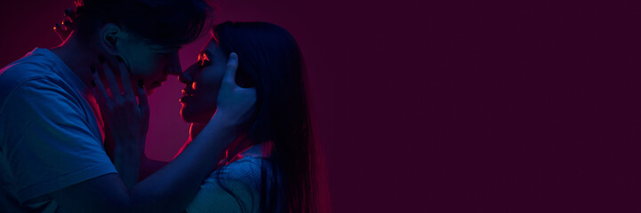 Portrait of young couple, boyfriend and girlfriend hugging and kissing, showing love against purple background in neon light. Concept of romance, love, relationship, passion, youth. Banner