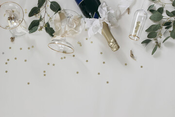 Happy New year party, celebration. Bottle champagne wine, drinking glasses, bows. Eucalyptus tree branches on white table background. Golden star confetti. Birthday anniversary flat lay, top view.