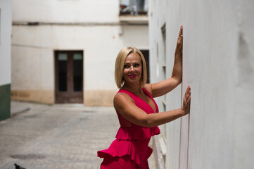 Beautiful blonde mature woman in an elegant red dress leaning against the white wall of a building...