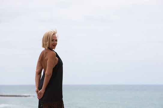 Mature, blonde and beautiful woman wears an elegant black dress and poses for the pictures happy and smiling while doing different poses. In the background the atlantic ocean in cadiz, spain.