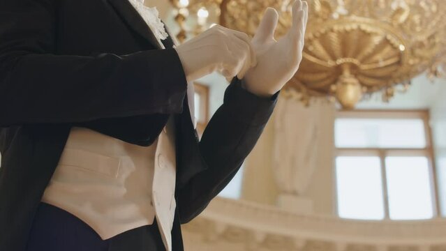 Midsection of unrecognizable male debutante adjusting tuxedo, vest and gloves while standing in Regency style ballroom with expensive gold chandelier hanging above