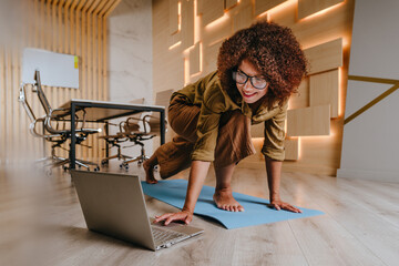 Woman practicing yoga at office using online lesson standing on a mat - obrazy, fototapety, plakaty