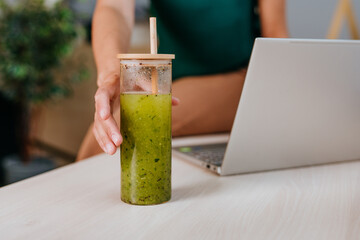 Close-up of woman drinking green smoothie with bamboo straw while using laptop