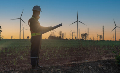 Asian male engineer wearing a hard hat stands in a turbine field holding blueprints, inspecting the operation of a wind power generating station to produce electricity, working until sunset.