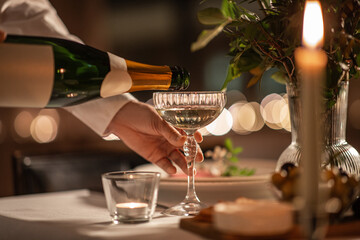 holidays, dinner party and celebration concept - close up of hand pouring champagne from bottle to glass on table at home or restaurant - obrazy, fototapety, plakaty