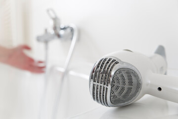 Hairdryer lying on a bathtub with hand and flowing water from a faucet in the background
