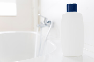 White bottle of shampoo standing on a bathtub in a bright bathroom