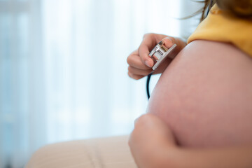 Asian pregnant woman listening to baby heartbeat with stethoscope at home