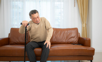 With knee joint pain, a senior guy sits on a sofa with a walking stick in his living room at home.