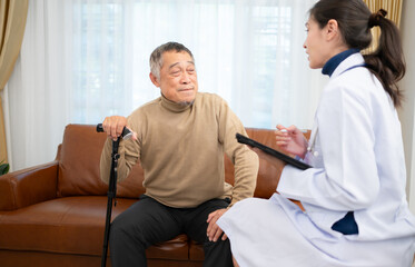 Doctor examines and treats an elderly man seated on a sofa with a cane who is suffering from knee joint pain.