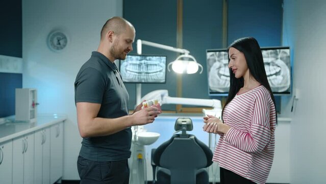 Amiable young dental expert in dark grey T-shirt and trousers, attractive young female patient standing in modern illuminated dentist office, talking. High quality 4k footage