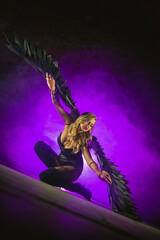 Obraz premium Beautiful girl an angel with the black wings posing in the smoke on dark background in the purple light. Angel of the night concept.