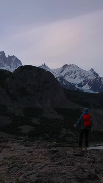Traveler Man with Backpack and Trekking Poles Looking at Mount Fitz Roy and Cerro Torre. El Chalten. Hills and Snow-Capped Mountains. Andes, Patagonia, Argentina. Aerial View. Orbiting. Vertical Video
