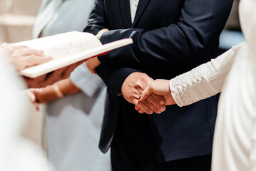Detalle de una pareja de novios agarrándose de las manos en el altar durante la ceremonia de casamiento frente al sacerdote. Concepto del sacramento del matrimonio en la iglesia Católica - obrazy, fototapety, plakaty