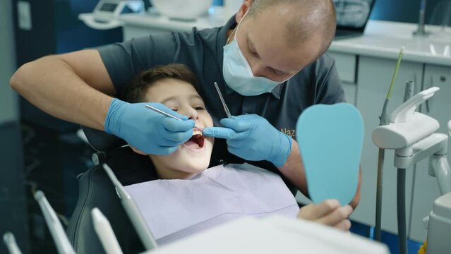 Calm boy sitting in dentist chair opening mouth wide while dental expert in latex gloves and medical mask checking teeth with tooth scraper, concave mirror. High quality 4k footage