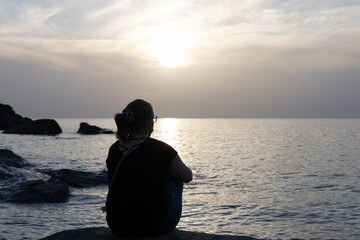 Back view of a lonely girl sitting by the aegean sea enjoying  the sunset scenery.