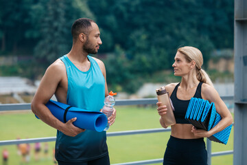 Couple wearing sportive clothes posing with yoga mat and water bottles before training on stadium.