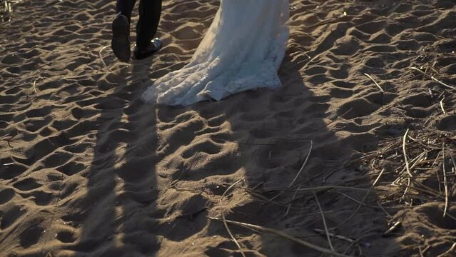 Young romantic couple in love walks at the beach in the evening sunset. Man and woman holding hands and hugs. Girl barefoot goes on sand. Bride and groom at their wedding in white dress and suit.