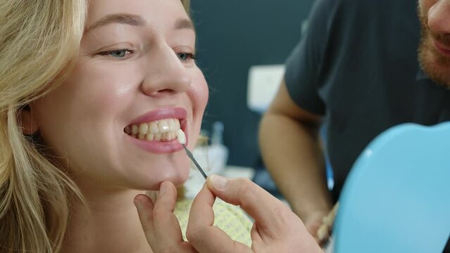 Nice woman with healthy bright skin looking in mirror at her teeth, male doctor applying sample of dental implant, light fully equipped dentist office. High quality 4k footage