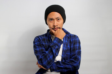 Pensive young Asian man, wearing a beanie hat and casual shirt, places his hand on his chin,...