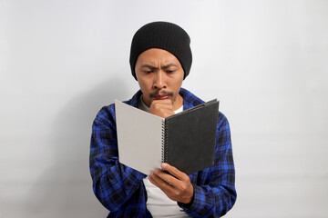 A serious young Asian student, dressed in a beanie hat and casual shirt, is deeply engrossed in...